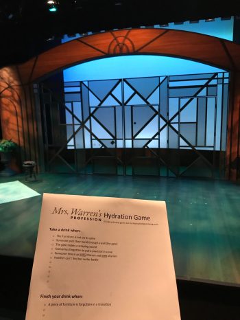 A piece of paper titled "Mrs Warren's Profession Tech Week Drinking Game" in front of a theatre set with a cyc lit blue and an art deco gate closed in front of it